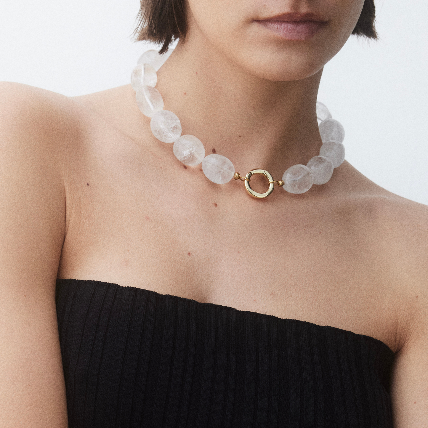 Woman wearing a chunky Clear Quartz necklace with round beads and a gold-plated brass clasp, styled with a black strapless top against a minimal white background.