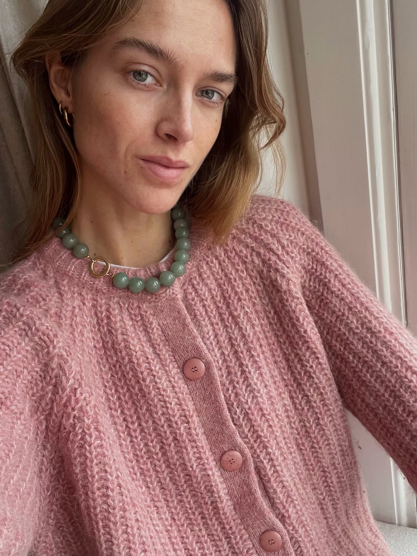 Close-up of a woman wearing a Green Aventurine beaded necklace with a brass clasp, styled with a soft pink textured knit cardigan — a cozy, feminine look with a crystal pop.