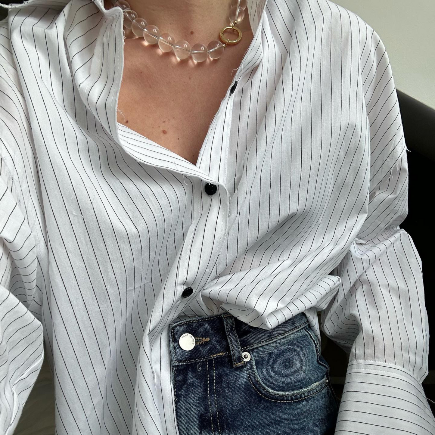 Close-up of a person wearing a clear quartz beaded necklace with a gold clasp, styled with a relaxed white pinstripe button-up shirt and high-waisted blue jeans.