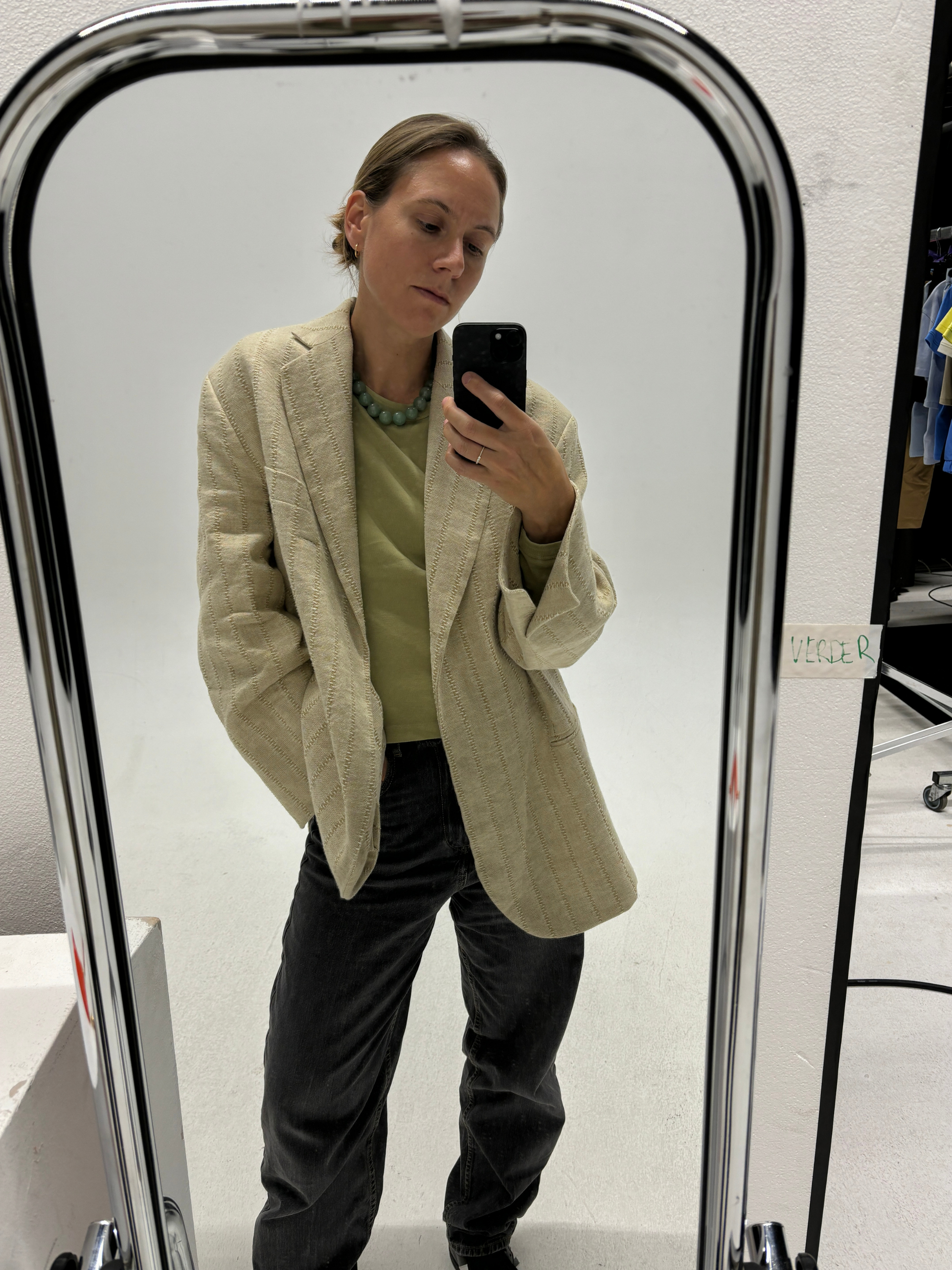 Woman taking a mirror selfie in a studio, wearing a Green Aventurine beaded necklace styled with a light green top, cream textured blazer, and black relaxed-fit jeans — a chic, casual look with a subtle crystal statement.