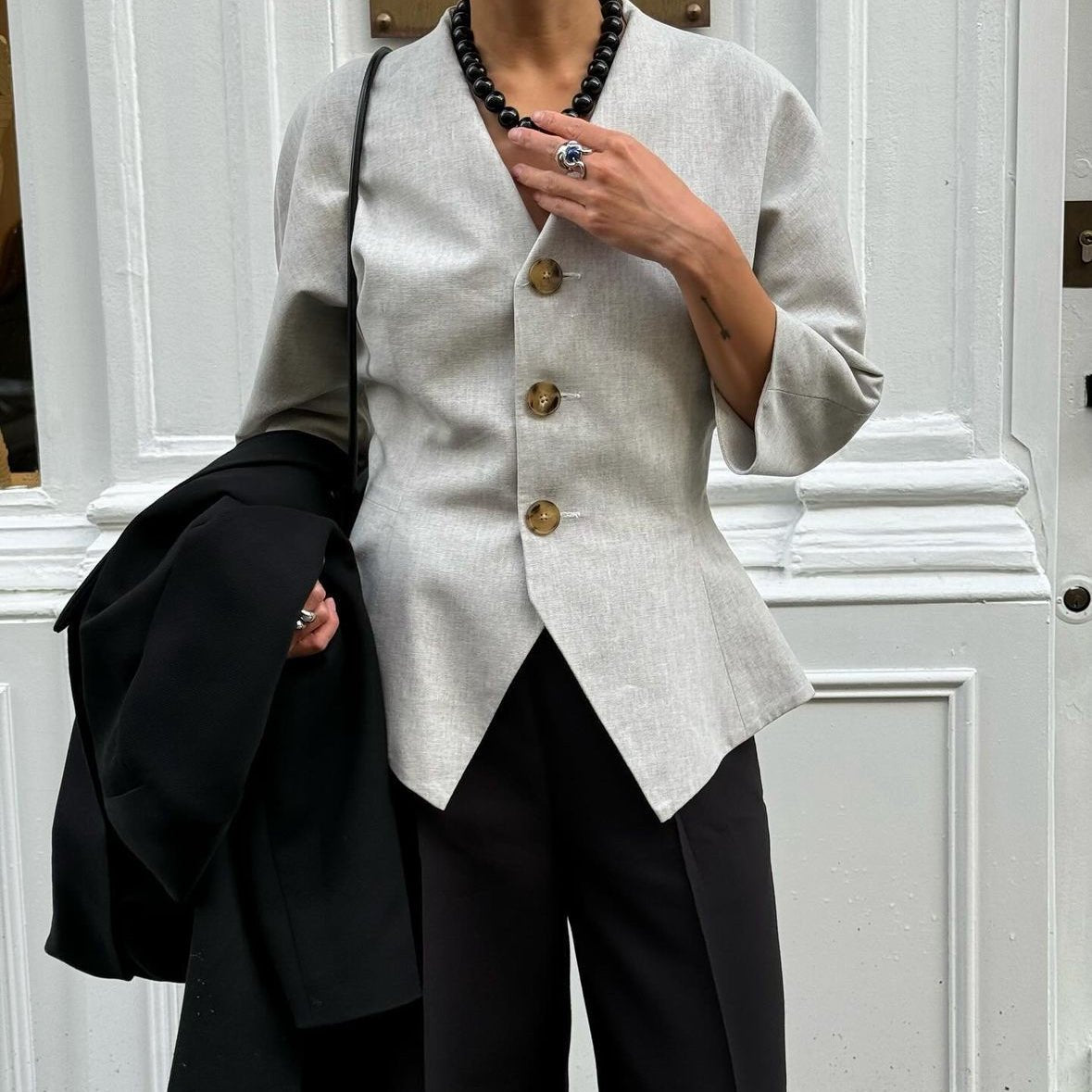 Street-style shot of a woman wearing the Grace Black Onyx Necklace with a tailored light grey blazer and black trousers, accessorized with a statement ring and black coat in hand.