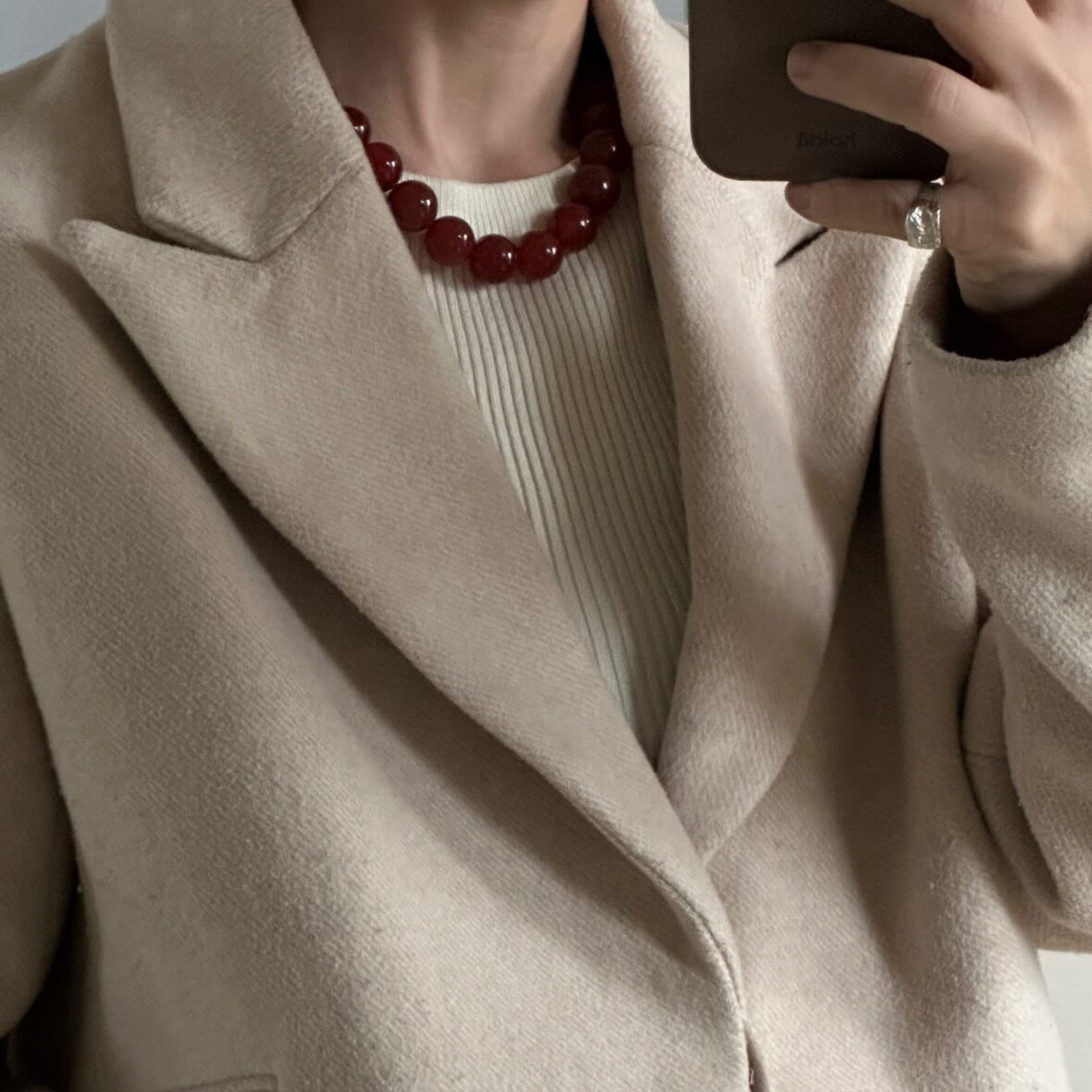 Close-up of a woman wearing a chunky Carnelian necklace styled with a ribbed cream top and beige wool coat, holding a phone.