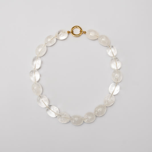 Overhead view of the Ignite Clear Quartz necklace featuring polished oval clear quartz beads, strung on transparent thread with a gold-plated brass spring ring clasp, displayed on a light grey background.