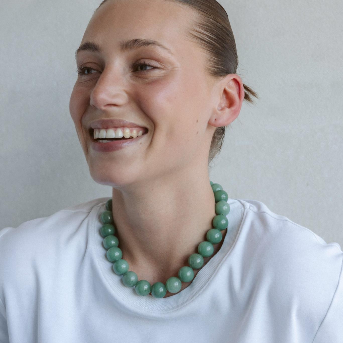 Close-up of a smiling woman wearing a bold green Aventurine beaded necklace styled with a white top, exuding joy and calm confidence.