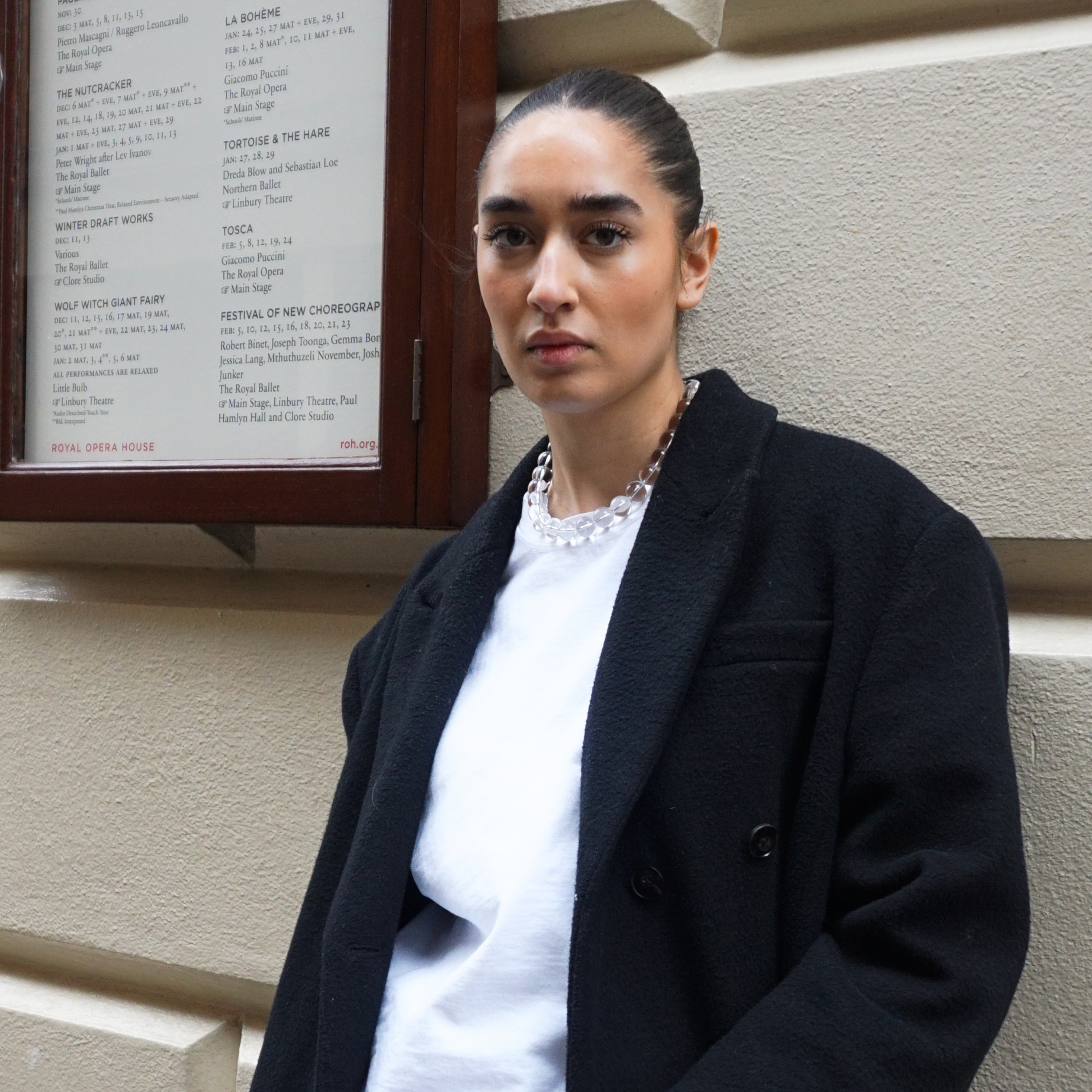 Woman standing in front of the Royal Opera House wearing a clear quartz bead necklace, a white t-shirt, and a black wool coat, looking directly at the camera with a confident expression.